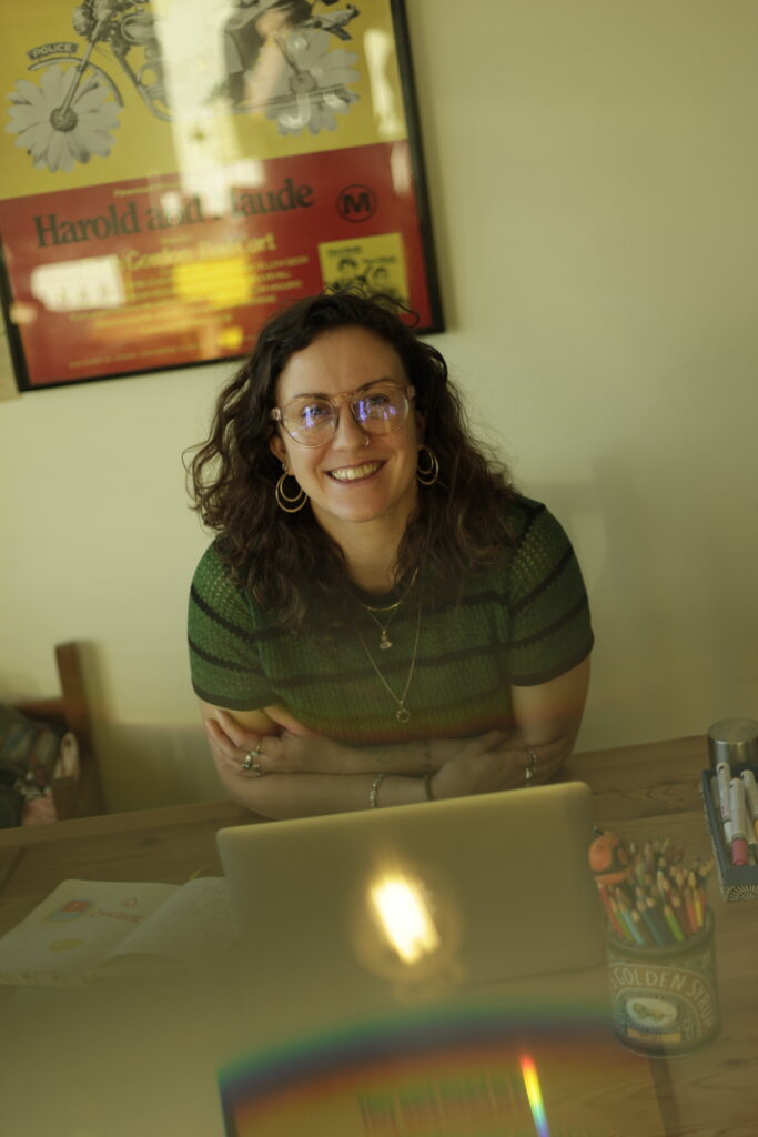 A woman with curly brown hair is sitting a table with her arms crossed and smiling at the camera. She has on open laptop in front of her.