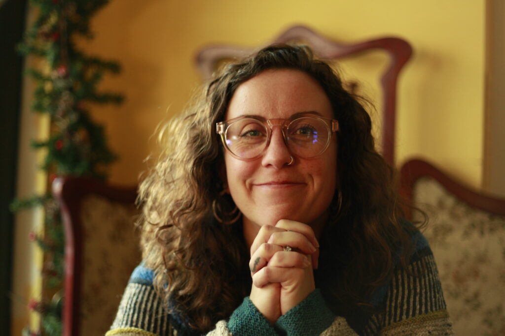 A woman with curly hair is smiling and look straight into the camera. She is resting her chin on her hands.
